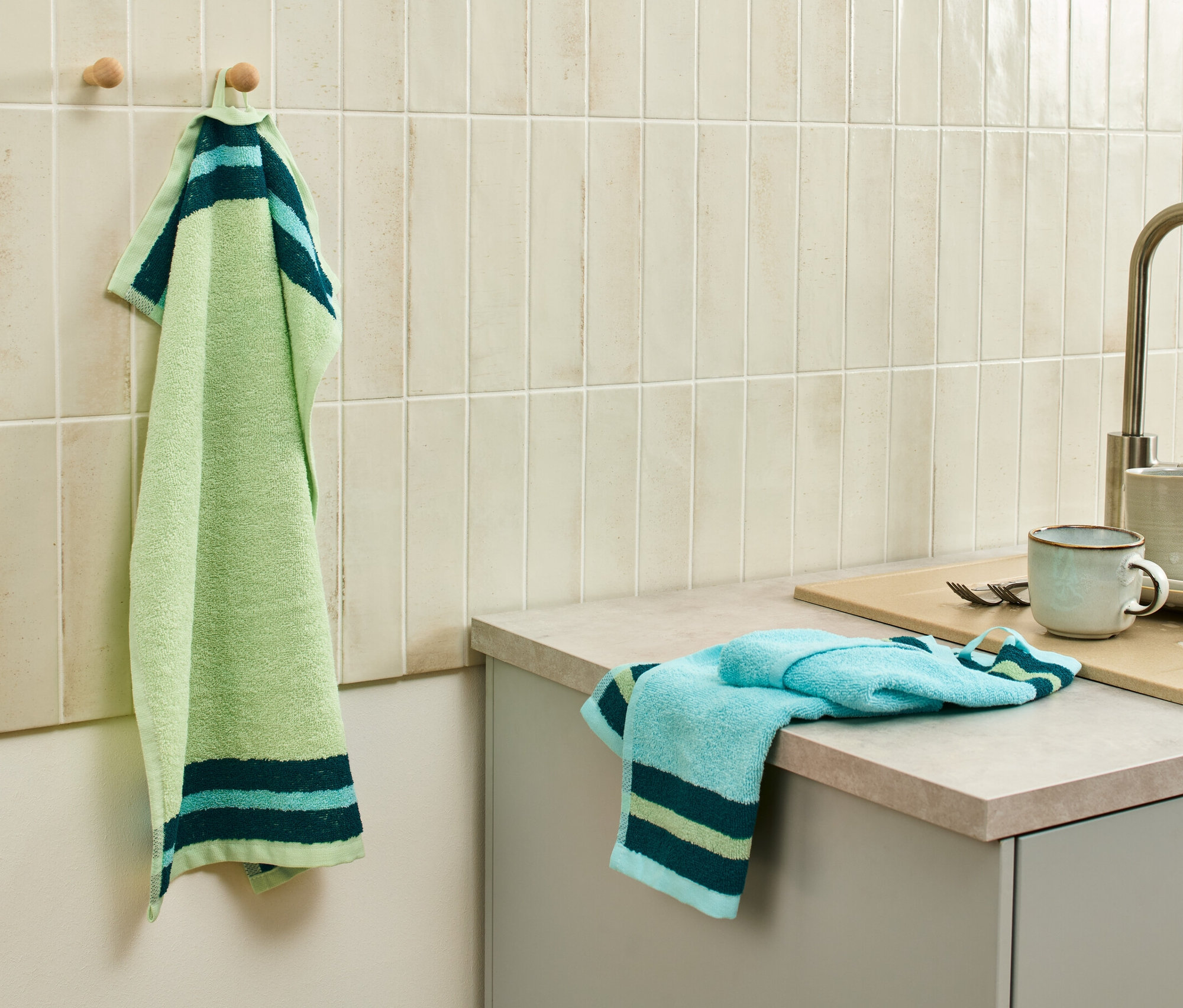 Egy zöld-kék konyharuha lóg egy akasztón a mosogató mellett, további 2 konyhai kéztörlő van a pulton.