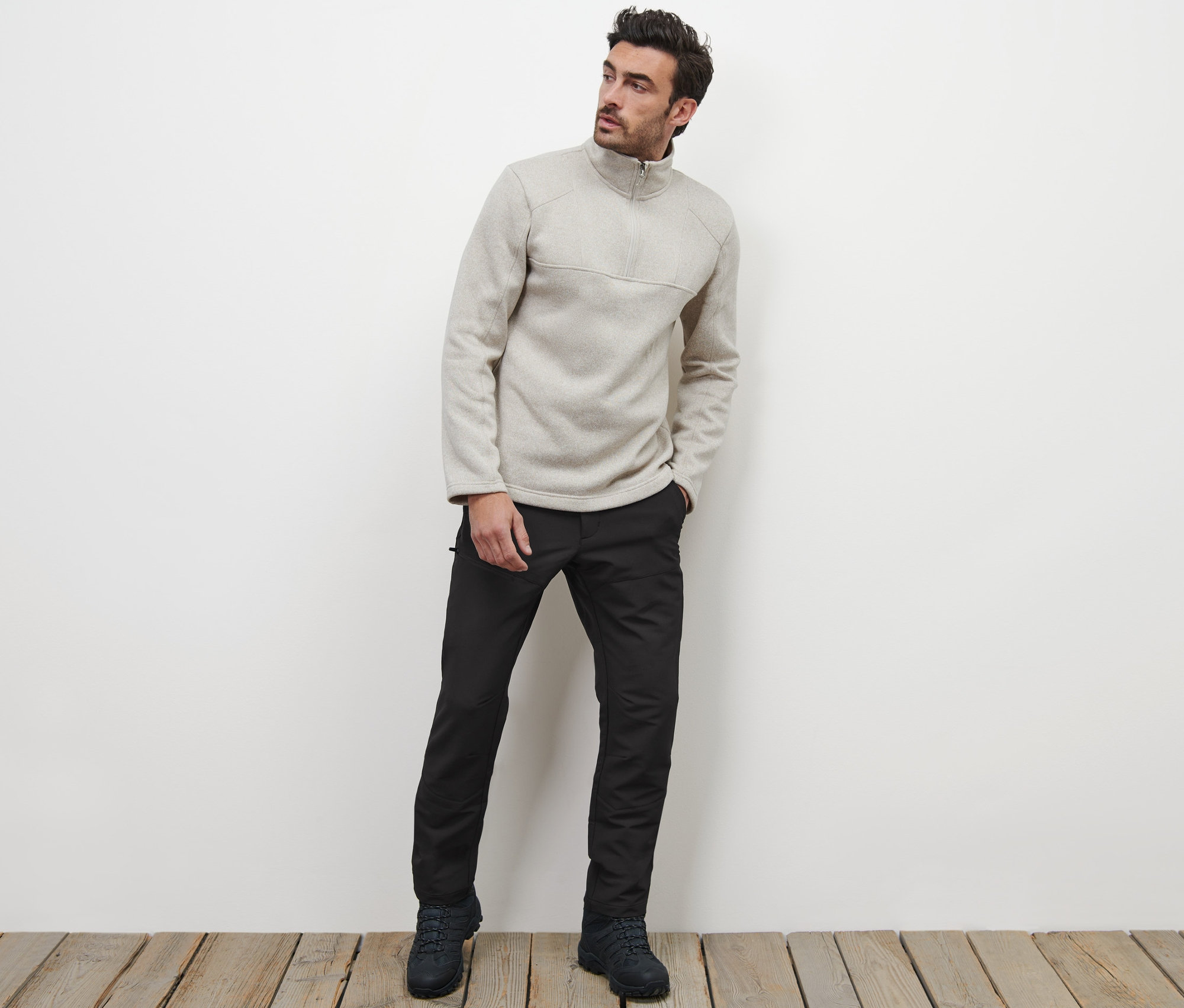 Férfi pózol fapadlón, termonadrágot és cipzáras pulóvert visel.