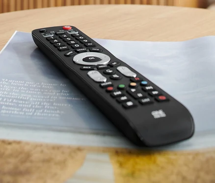 Egy fekete 4 az 1-ben univerzális távirányító fekszik egy asztalon egy pohár víz és egy magazin mellett.