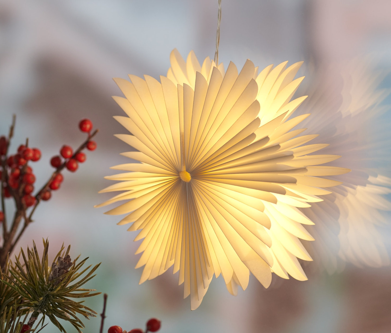 Ablak előtt függő, kicsi LED-es papírcsillag, mellette piros bogyós ágak.