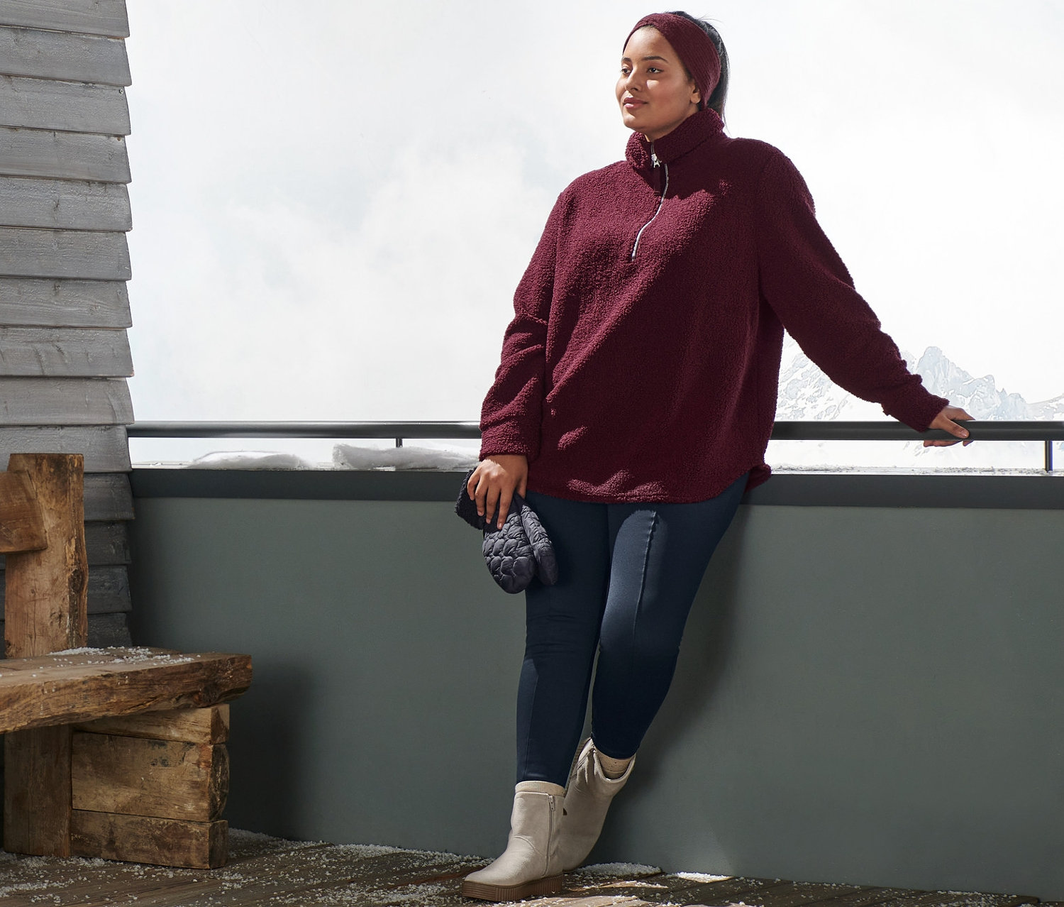 Egy nő egy korlátnak dőlve női oversize plüsspulóvert, bordó kötött fejpántot, sötétkék termoleggingst és taupe velúrbőr csizmát visel. A kezében sötétkék steppelt egyujjas kesztyű van.