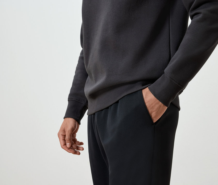 Egy férfi kezét a fekete melegítőnadrágja zsebében tartja, és sötétszürke pulóvert visel.