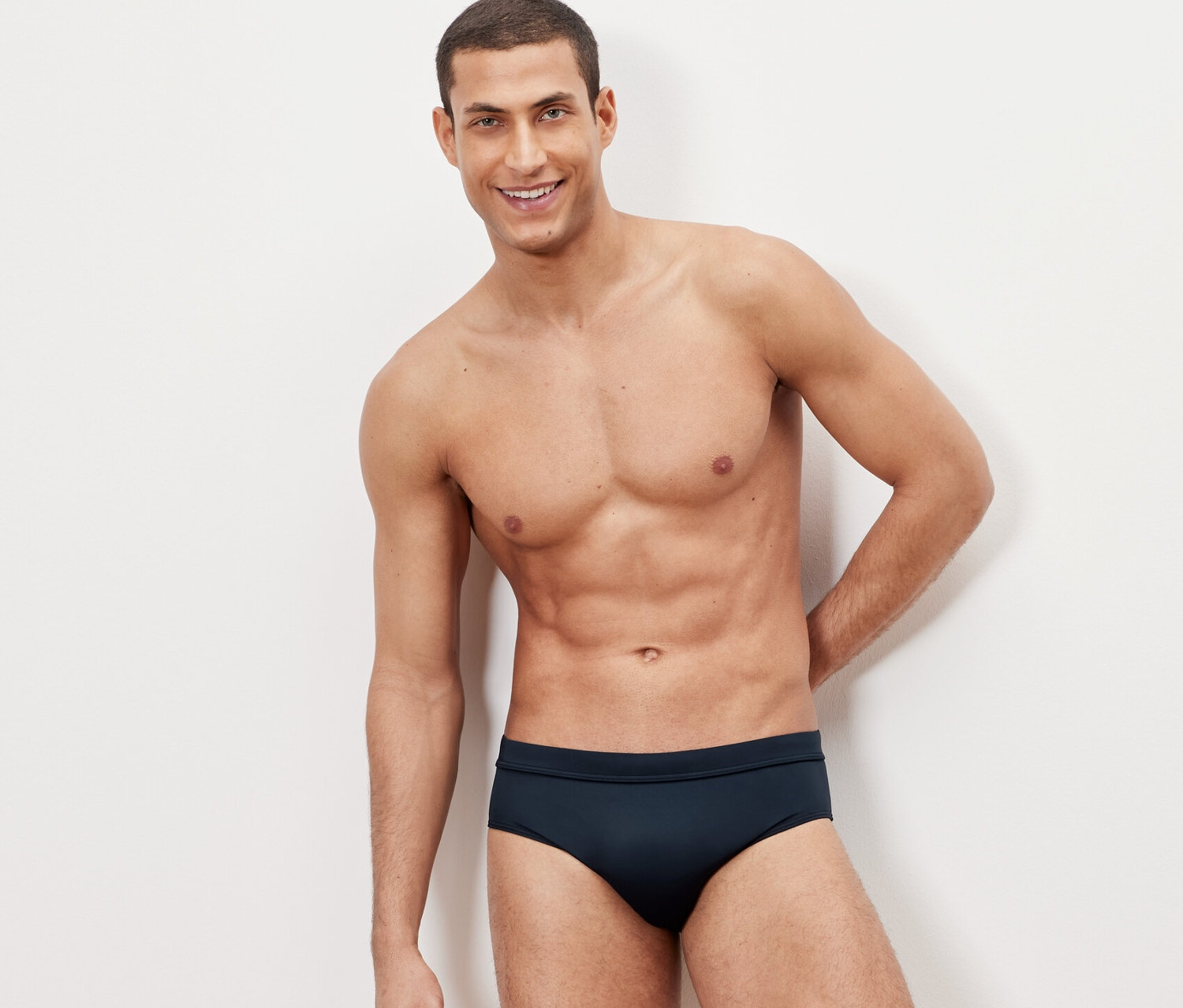 Férfi pózol fehér háttér előtt sötétkék férfi fürdőnadrágban.