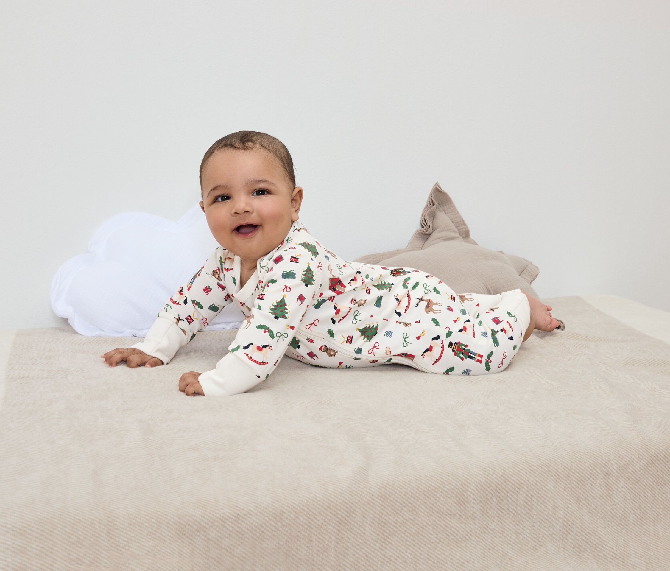 Egy mosolygó csecsemő hason fekszik egy karácsonyi mintás takarón, egy fehér felhőpárna és egy barna csillagpárna mellett.