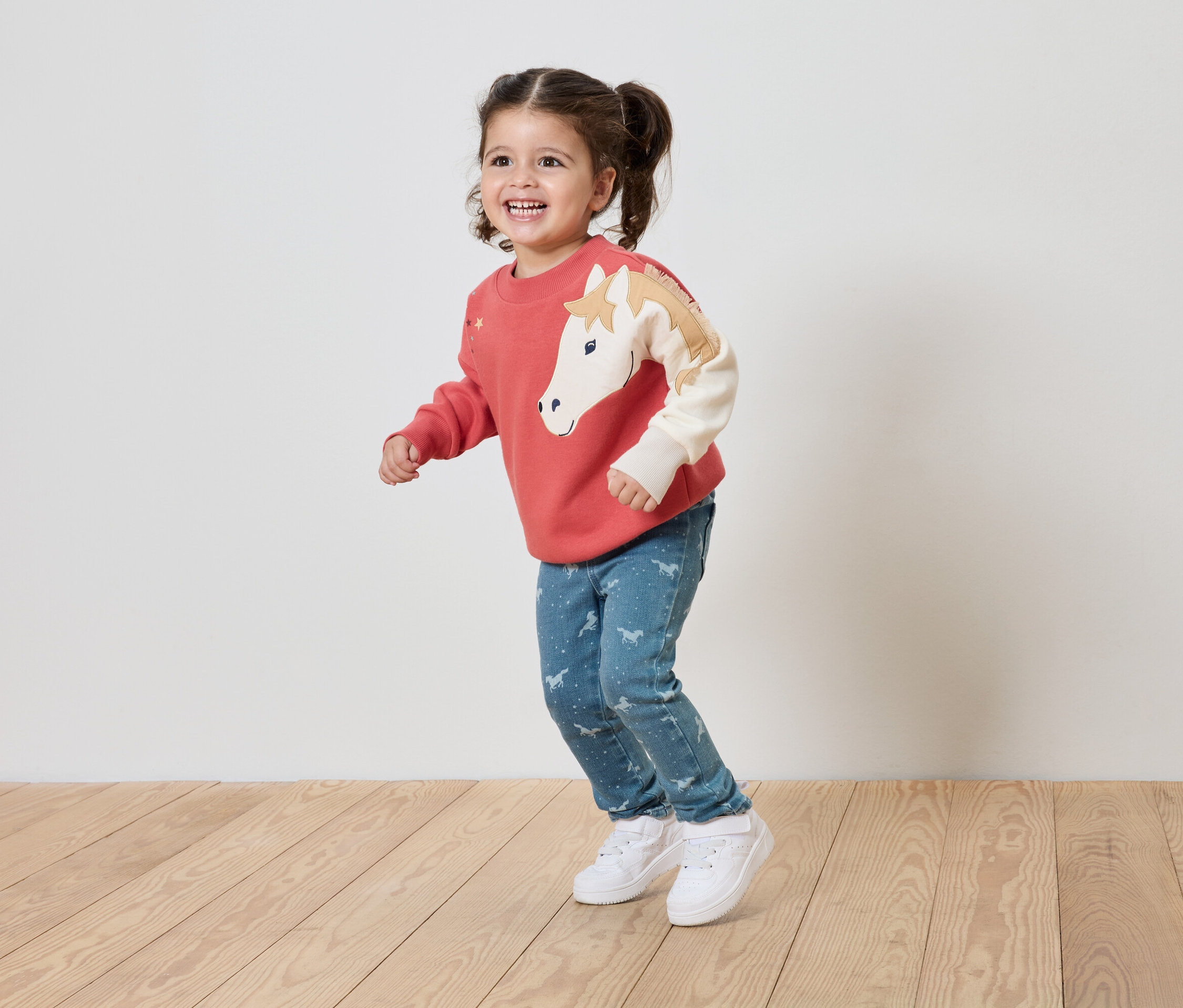 Mosolygó kislány korall színű, lovas pulóverben és kék, mintás farmerben áll a fapadlón.