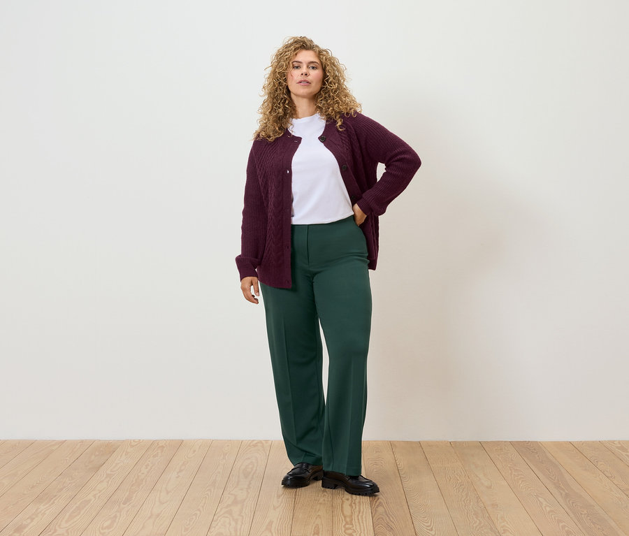 Egy nő áll, kezét a csípőjén tartja, bordó kardigánt, fehér pólót és zöld nadrágot visel.
