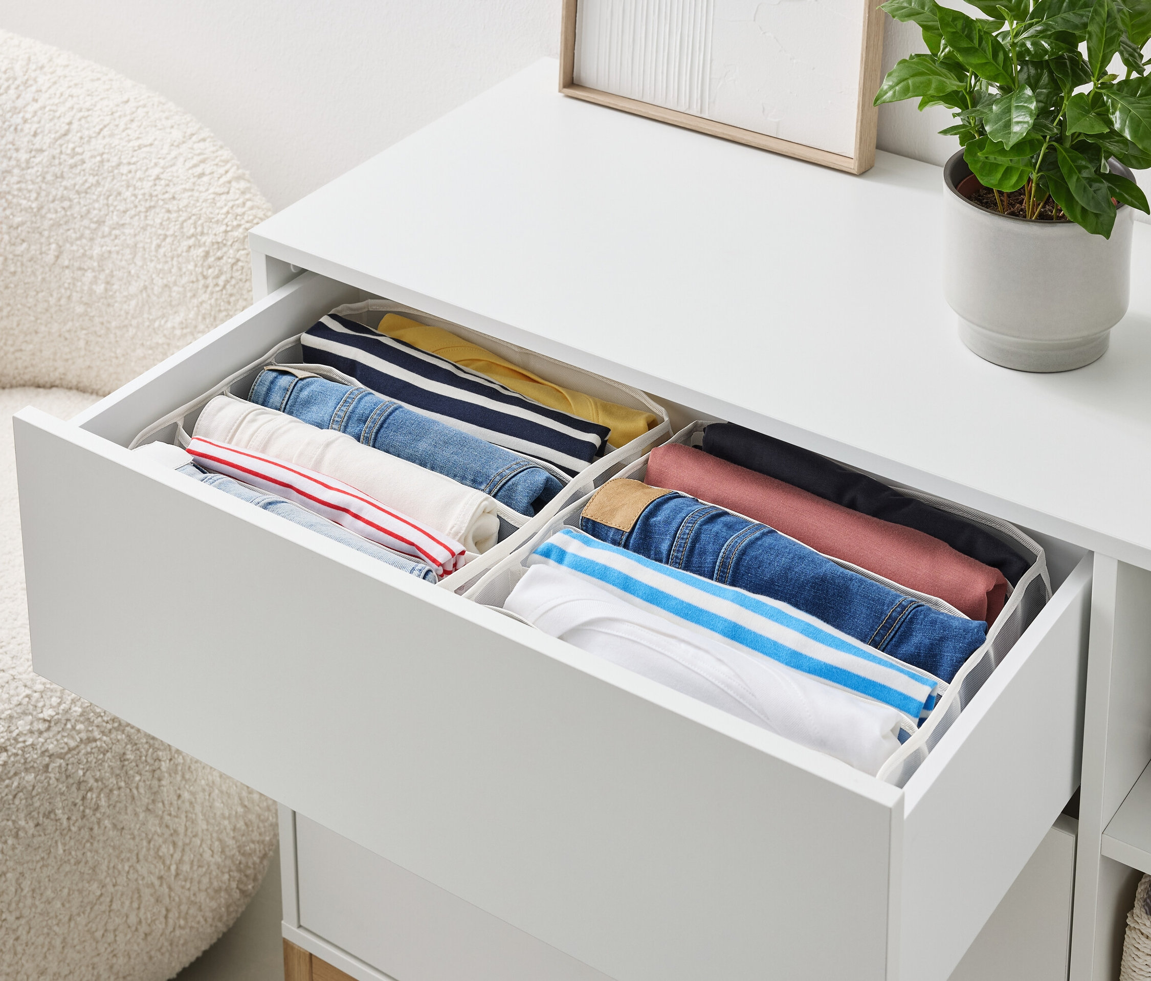 Egy fehér komód nyitott fiókjában rendezetten összehajtogatott ruhák találhatók tárolókban.