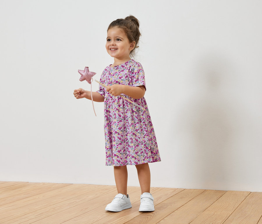 Mosolygó kislány csillagpálcát tart, virágos ruhát és tornacipőt visel.