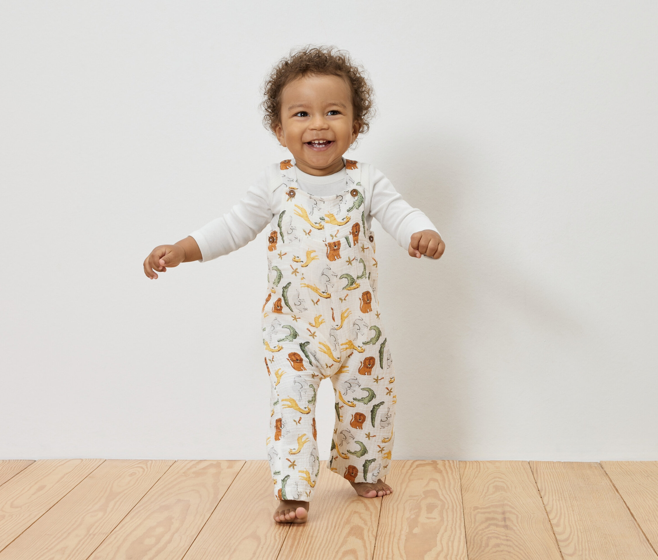 Egy mosolygó, göndör hajú kisgyermek áll egy fapadlón, hosszú ujjú fehér blúzt visel egy állatmintás fehér overall alatt.