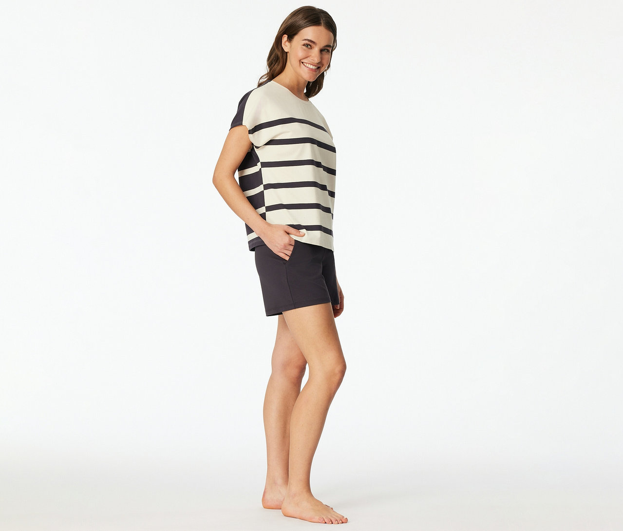 Nő visel egy SCHIESSER rövidnadrágos pizsamát csíkos felsővel és sötét rövidnadrággal, mosolyogva, kezét a zsebében tartva.