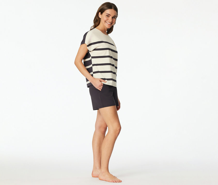 Nő visel egy SCHIESSER rövidnadrágos pizsamát csíkos felsővel és sötét rövidnadrággal, mosolyogva, kezét a zsebében tartva.