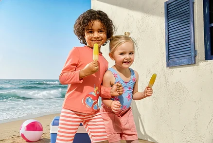 Két gyerek fagylaltozik a strandon. A lány Kinder-Badeanzug, Seesterne fürdőruhát és Kinder-Jeansshorts rövidnadrágot visel. A fiú Kinder-Kombi mit UV-Schutzfaktor 50+, Krake szettet visel.