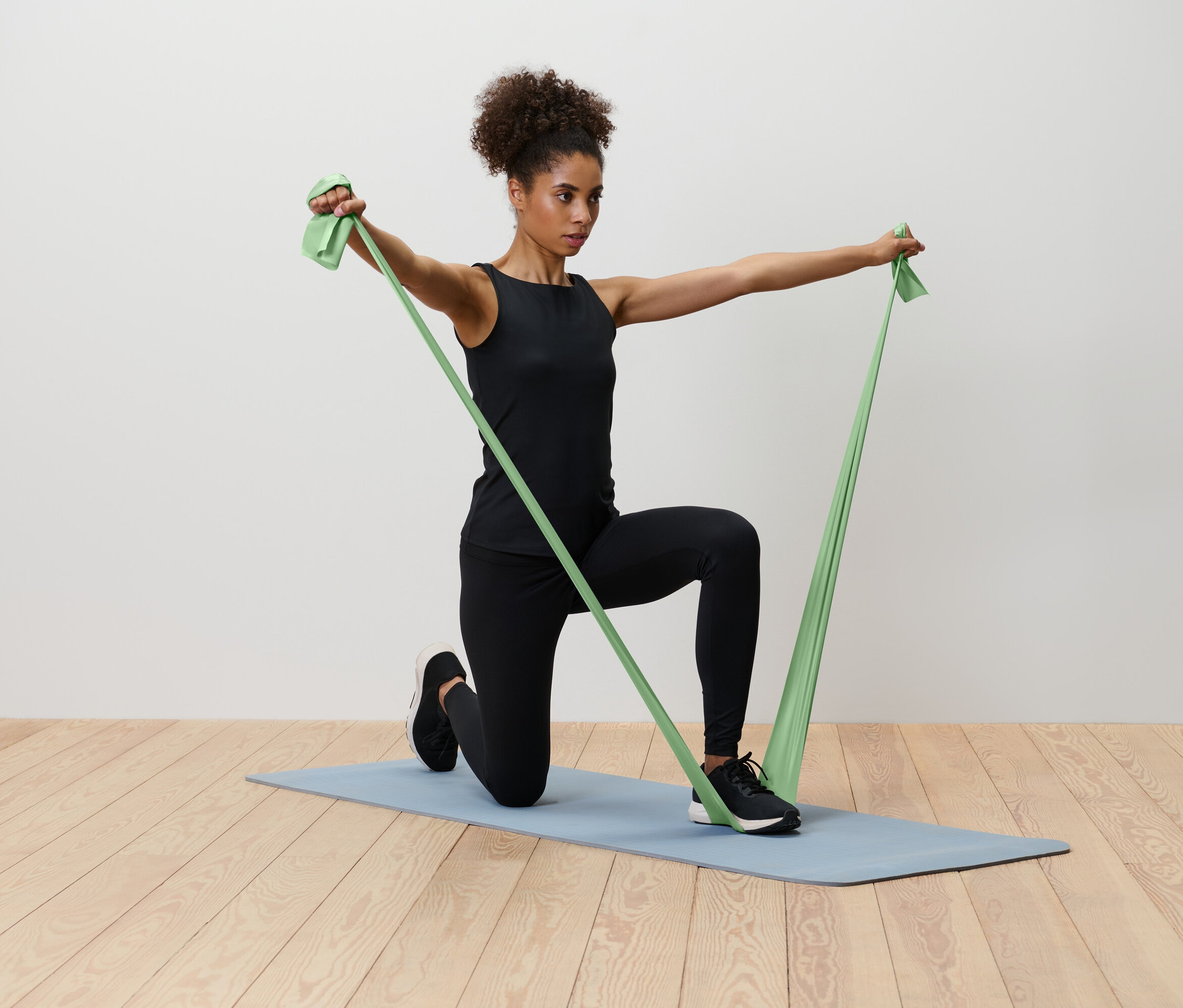 Egy nő térdel egy kék jógaszőnyegen, és egy zöld ellenállási szalagot nyújt.