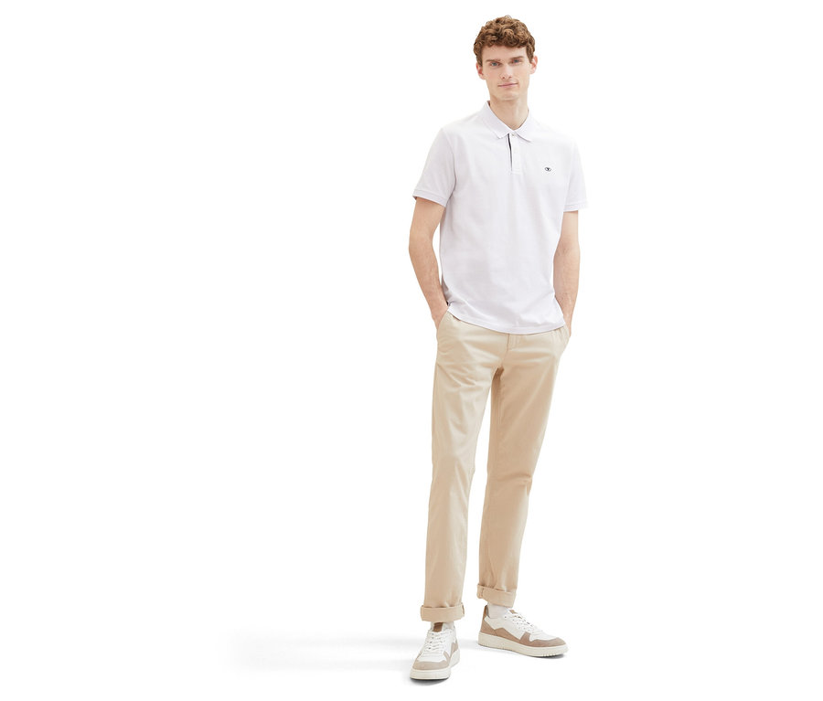 Férfi bézs nadrágban és fehér Tom Tailor piké pólóingben. Kezeit a zsebébe téve áll fehér háttér előtt.