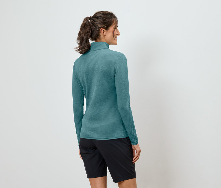 A lófarokba kötött hajú nő türkizkék, hosszú ujjú felsőt és fekete rövidnadrágot visel.