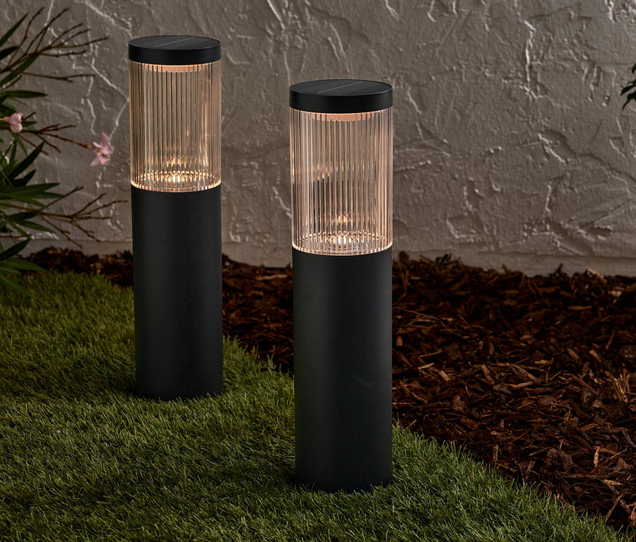 Két fekete szolárlámpa áll a gyepen egy fal előtt.