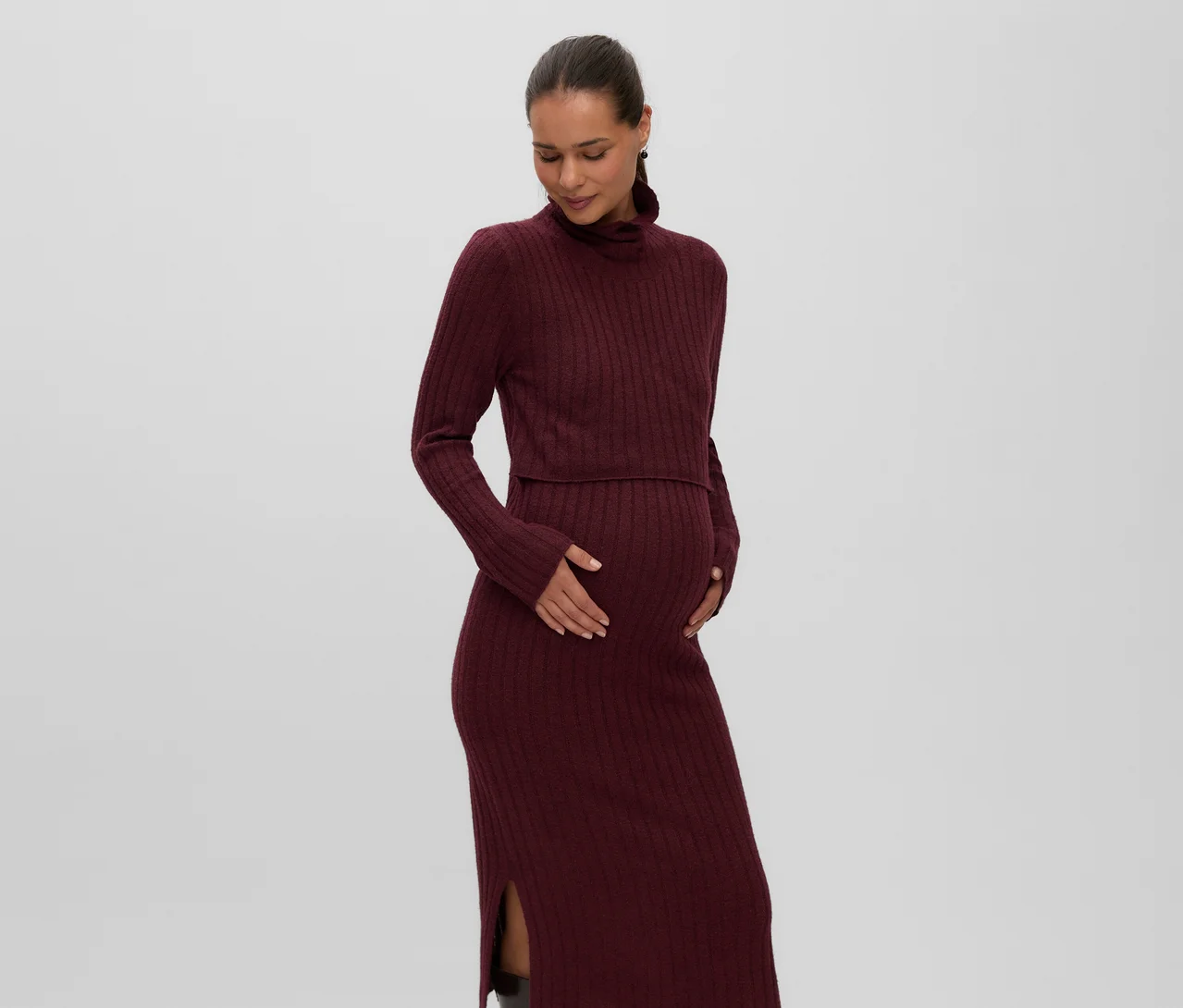 Terhes nő bordó színű, bordázott MAMALICIOUS »SUNIVA« midi kötött ruhát visel magas nyakkal és oldalsó sliccel, kezét a hasán tartja.