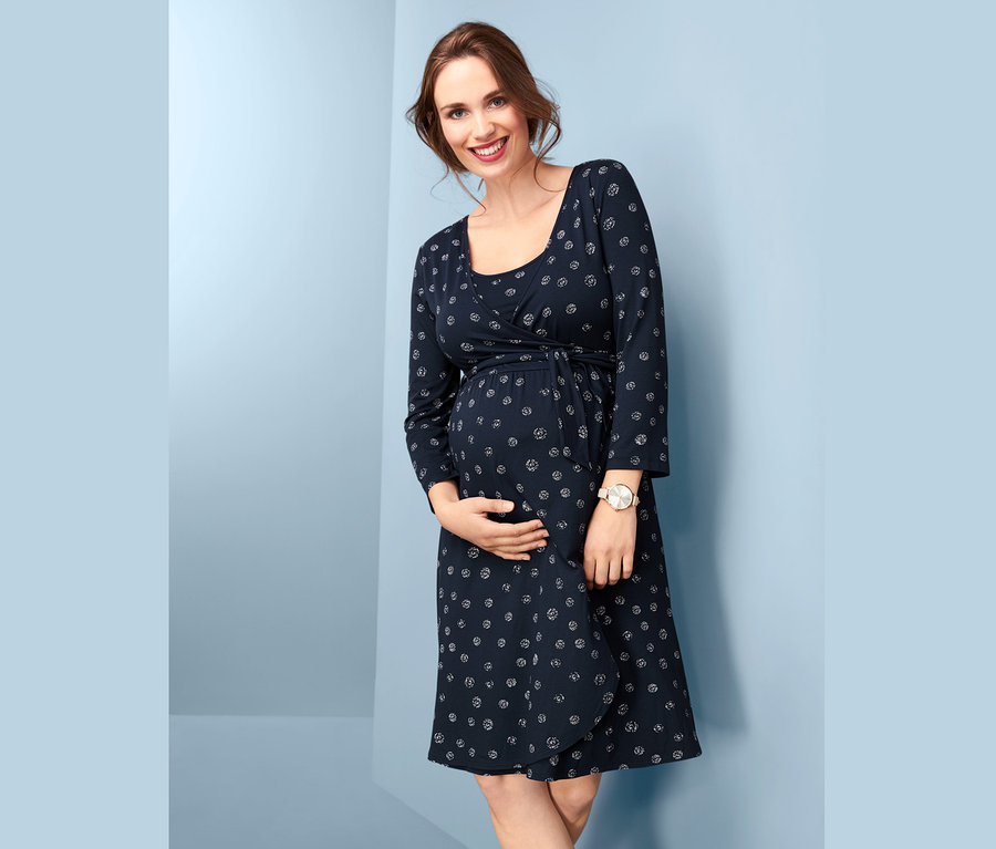 Egy mosolygó, terhes nő a hasát tartja, és egy sötétkék, mintás kismama ruhát visel.