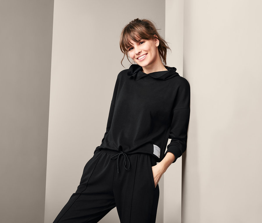 Mosolygó nő dől a falnak fekete női lezser felsőben és nadrágban.