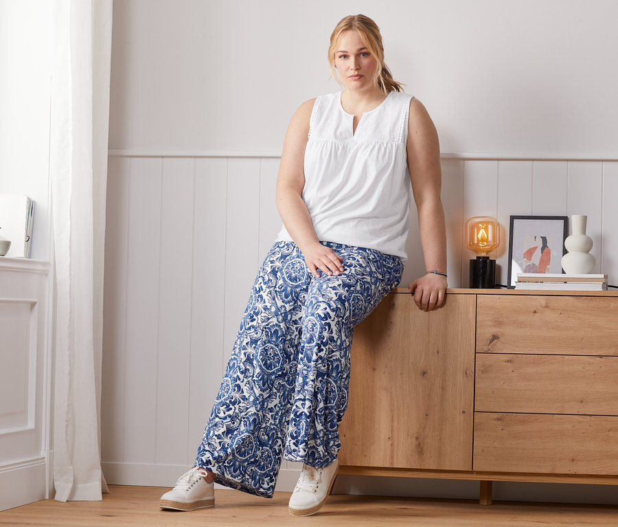 Nő ül a komódon, mintás, kék női bőszárú nadrágot és fehér, lyukhímzéssel díszített női topot visel.