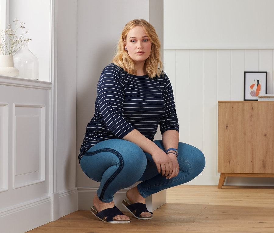 Nő kék jeggingsben és csíkos denevérujjú felsőben pózol.