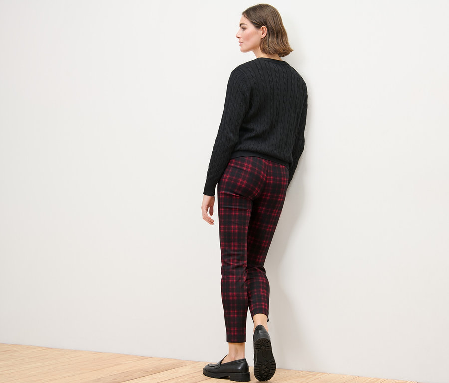 Egy nő áll háttal a kamerának, kockás sztreccsnadrágot, fekete pulóvert és fekete cipőt visel.