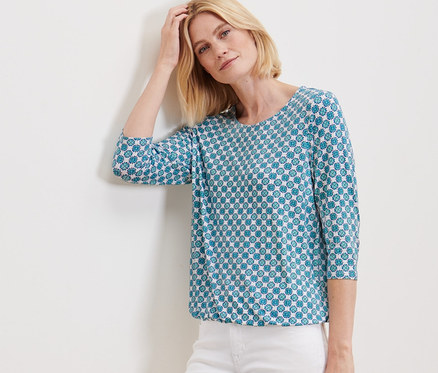 Nő a falnak támaszkodva, nyomott mintás női blúzban és fehér nadrágban pózol.