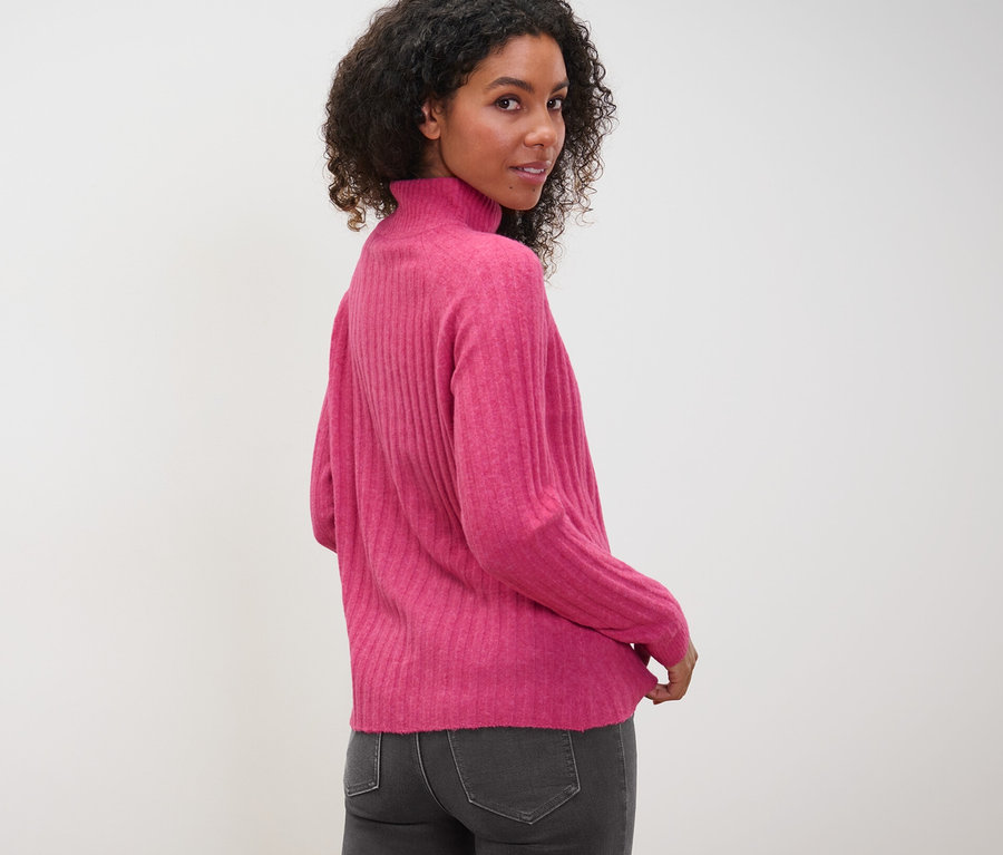 Nő néz a válla fölött, bordázott kötött garbót és szürke farmert visel.