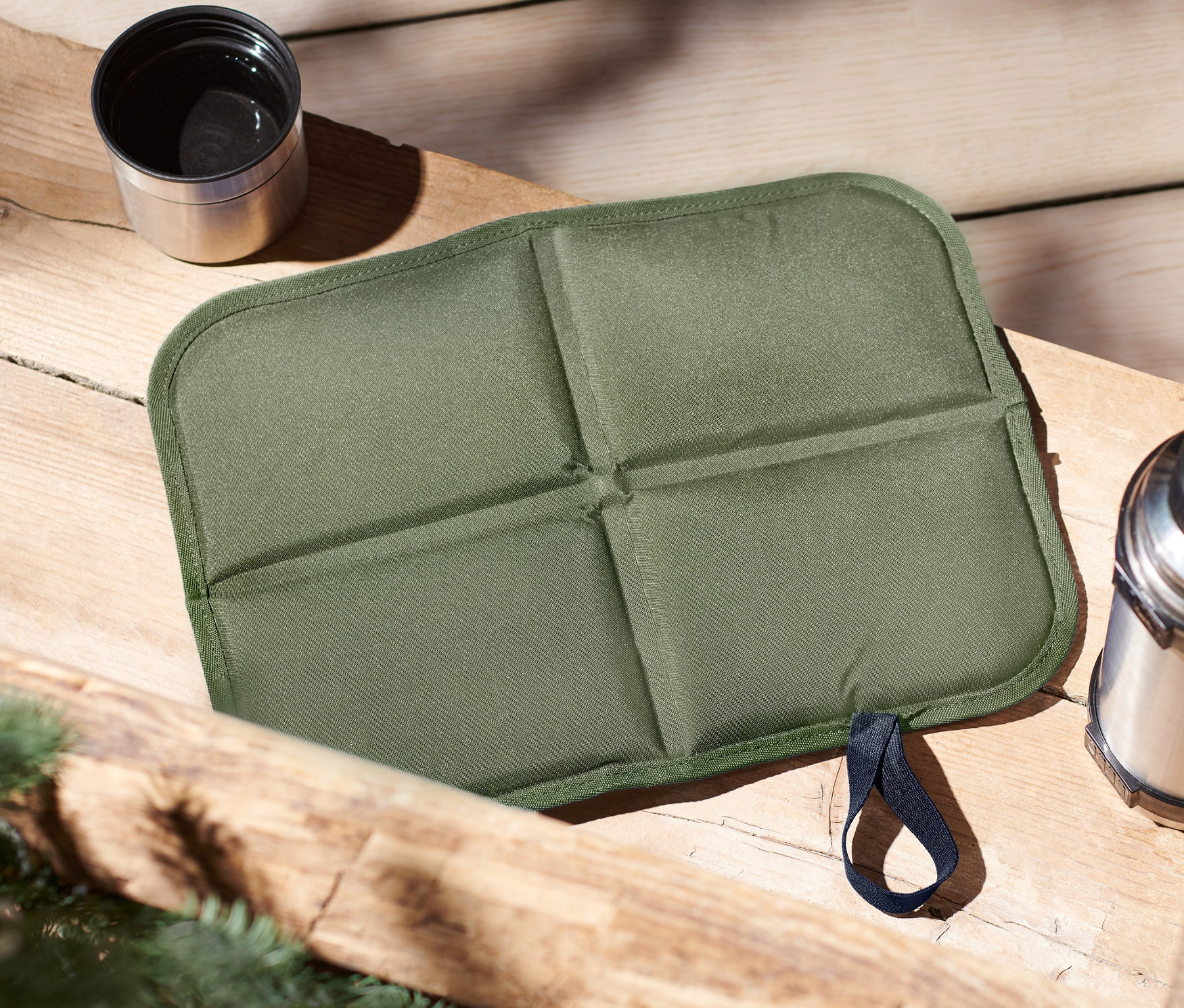 Egy fa asztalon egy zöld, összehajtható termo ülőpárna, egy termosz és egy csésze található.