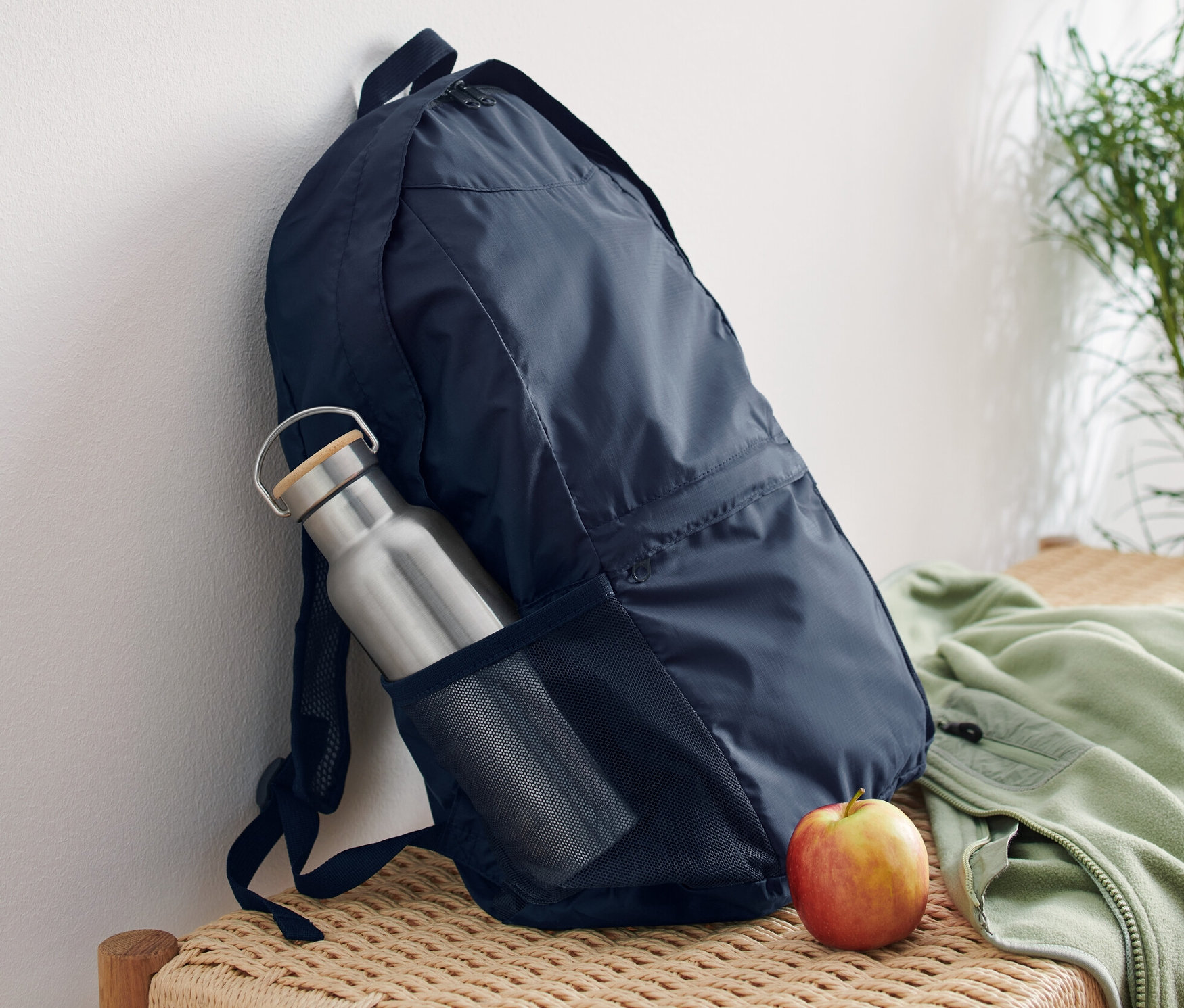 Egy sötétkék, összehajtható hátizsák támaszkodik egy fehér falnak egy vesszőpadon. A hátizsák oldalzsebében egy ezüst színű kulacs van, mellette pedig egy alma fekszik.