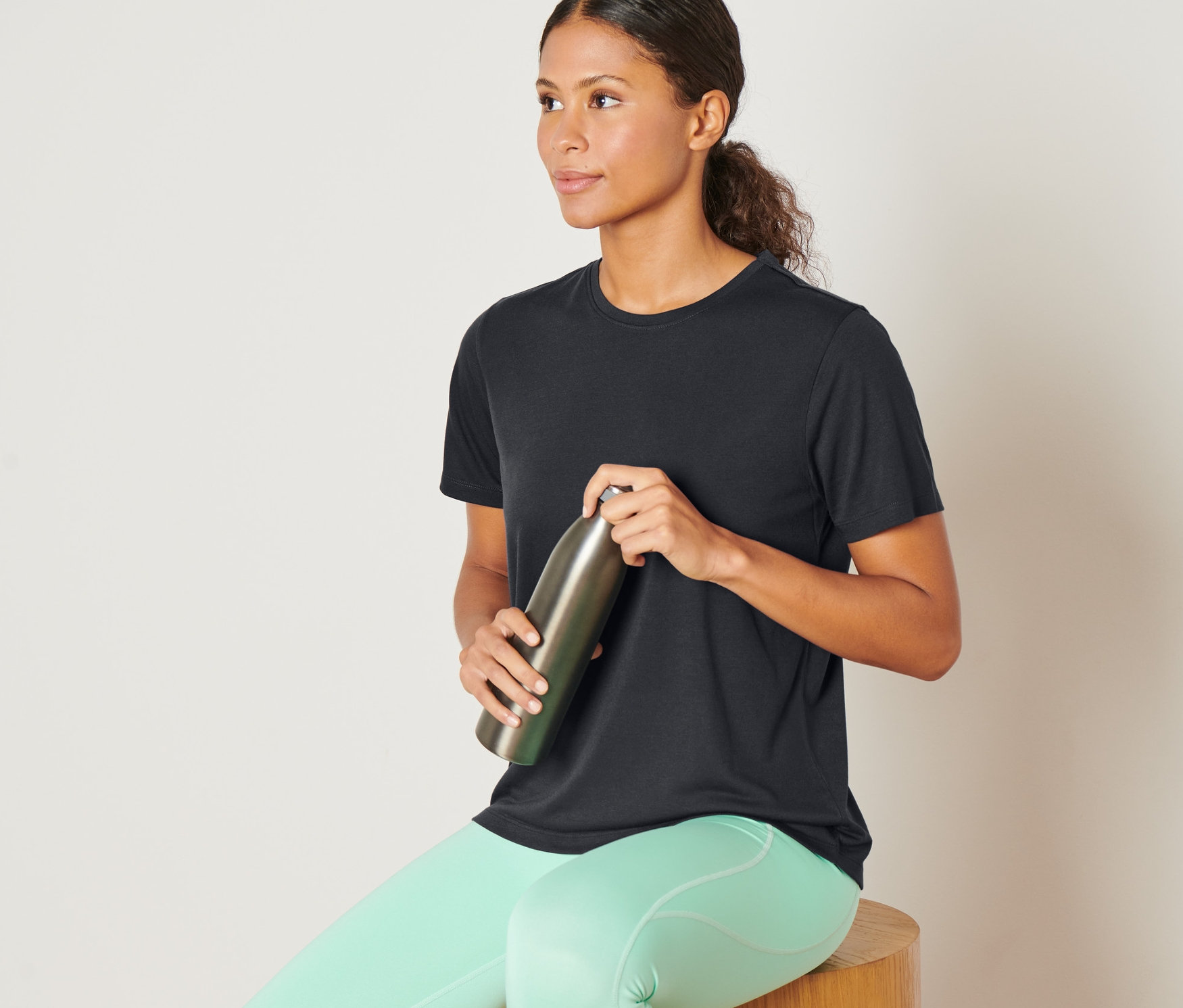 Nő ül egy fa ülőkén, antracit sportfelsőt visel, és egy fém palackot tart.