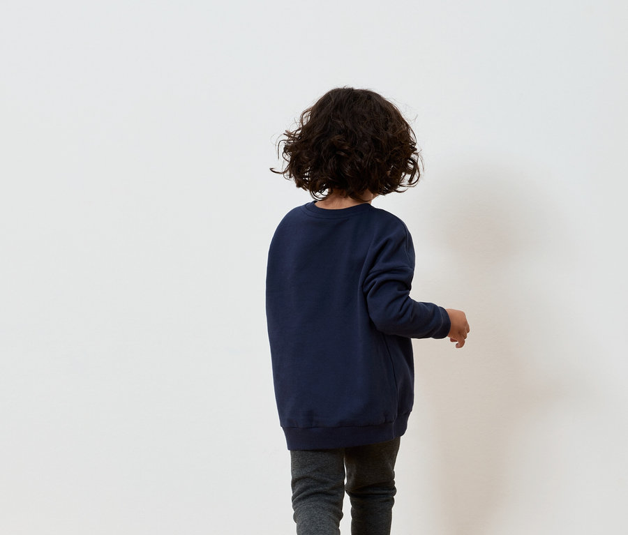 Egy barna göndör hajú fiú sötétkék pulóverben és szürke nadrágban áll háttal egy fehér falnak.