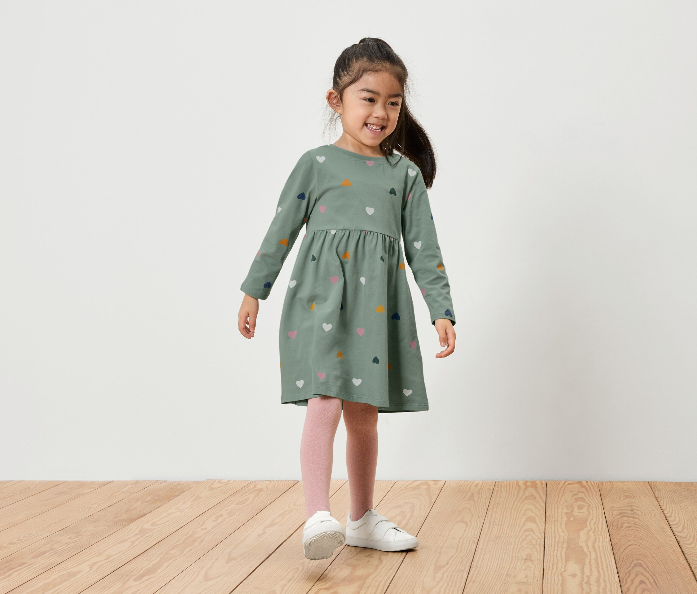 Egy mosolygó kislány áll egy fapadlón. Zöld, hosszú ujjú, szívecskés mintás ruhát, rózsaszín leggingset és fehér cipőt visel.