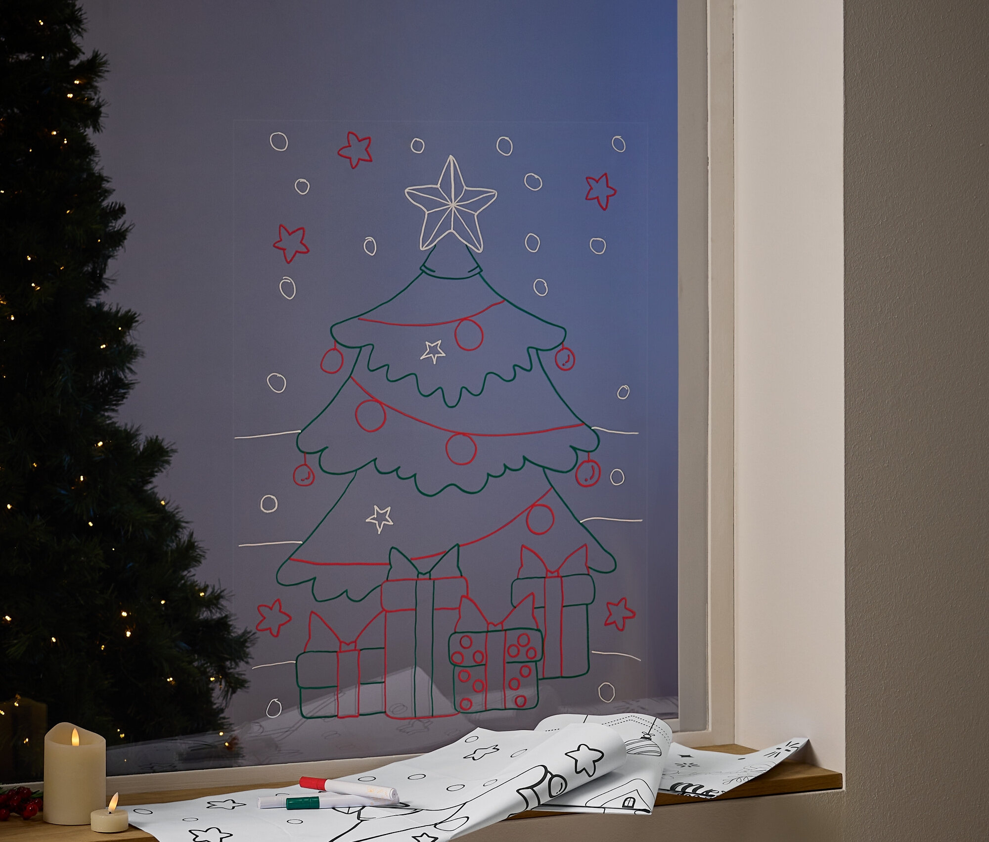 Egy ablakon karácsonyfa van rajzolva díszekkel és ajándékokkal. Az ablakpárkányon karácsonyi mintás papír és filctollak vannak.