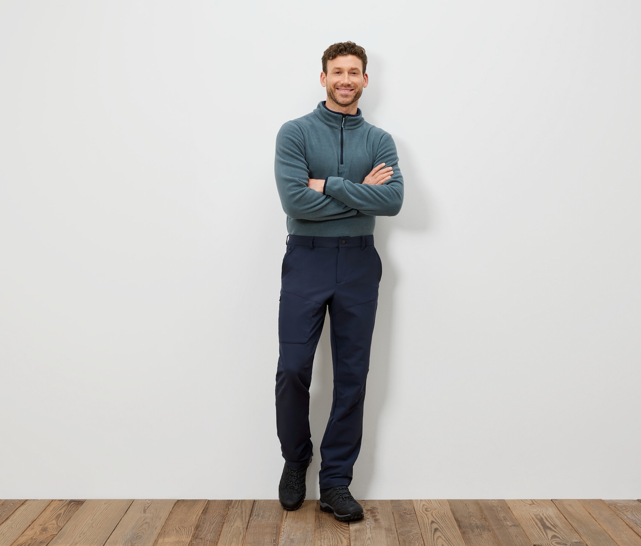 Egy férfi áll keresztbe tett karokkal, kékeszöld pulóvert és sötétkék nadrágot visel.