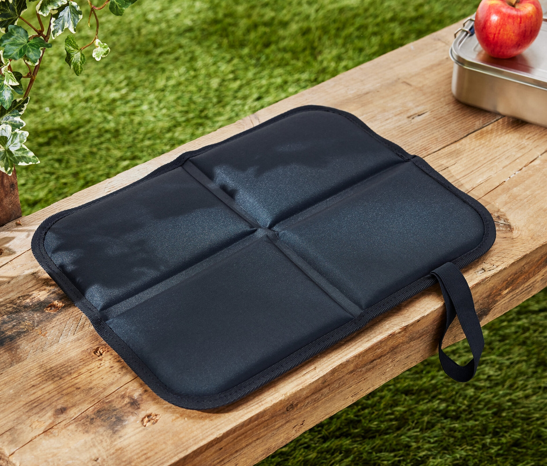 Egy összehajtható termo ülőpárna fekszik egy fa padon egy alma és egy uzsonnás doboz mellett.