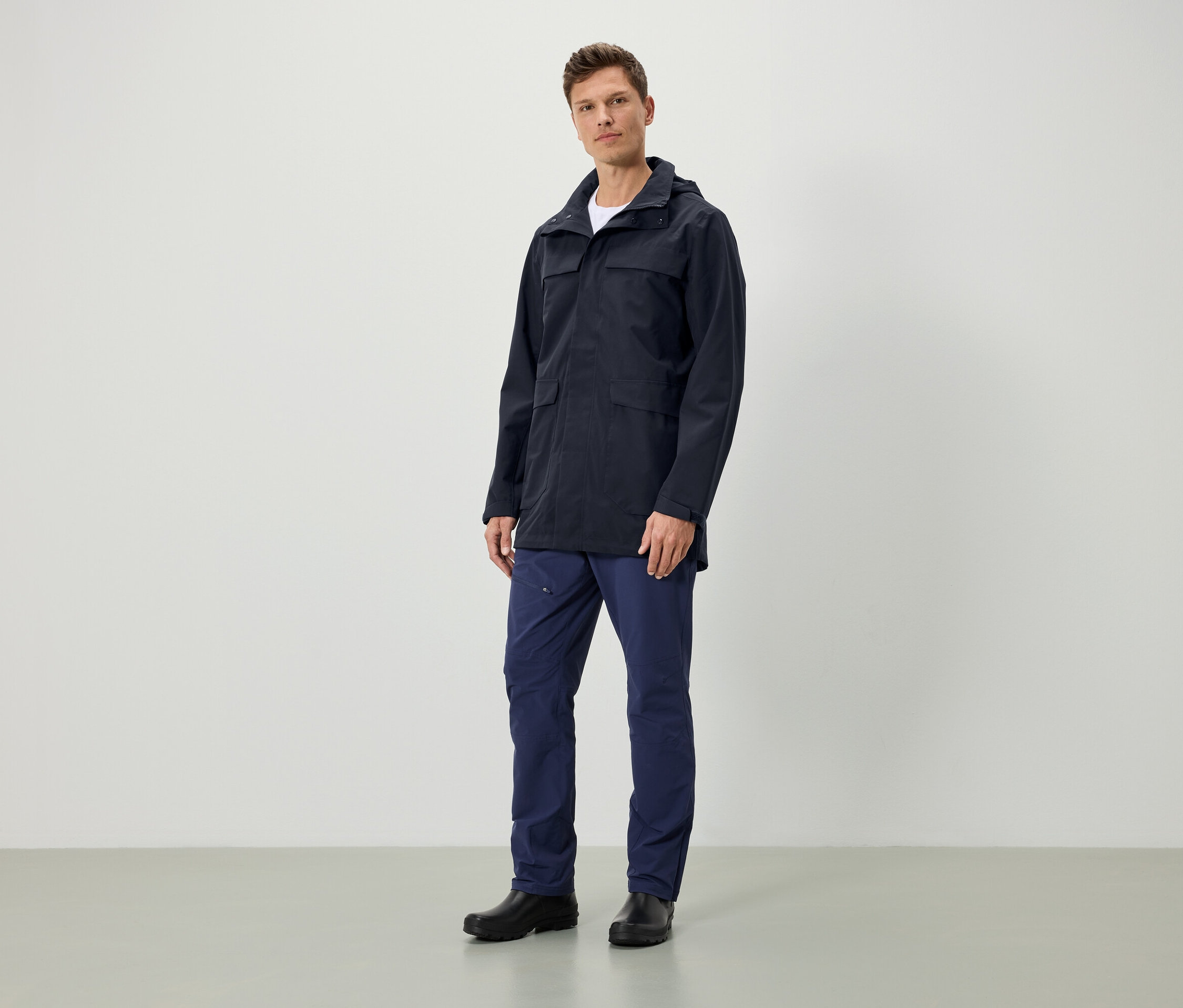 Egy férfi áll, sötétkék kapucnis kabátot, sötétkék nadrágot és fekete cipőt visel.