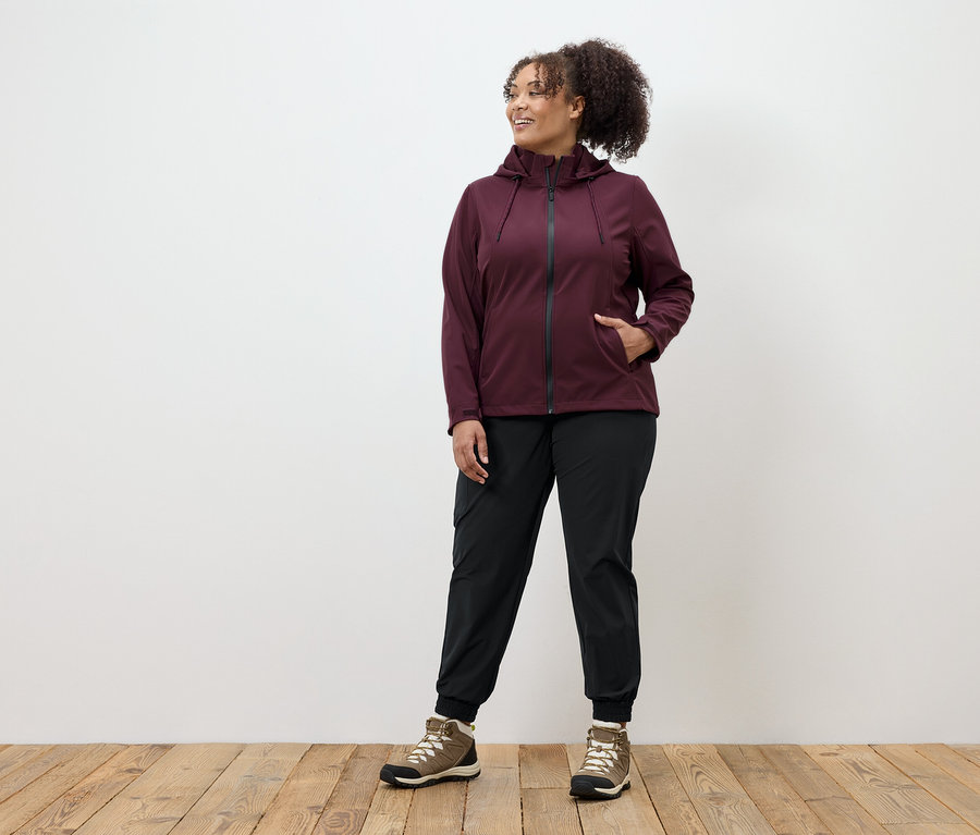 Egy nő bordó cipzáras kabátban, fekete nadrágban és barna cipőben áll egy fapadlón.