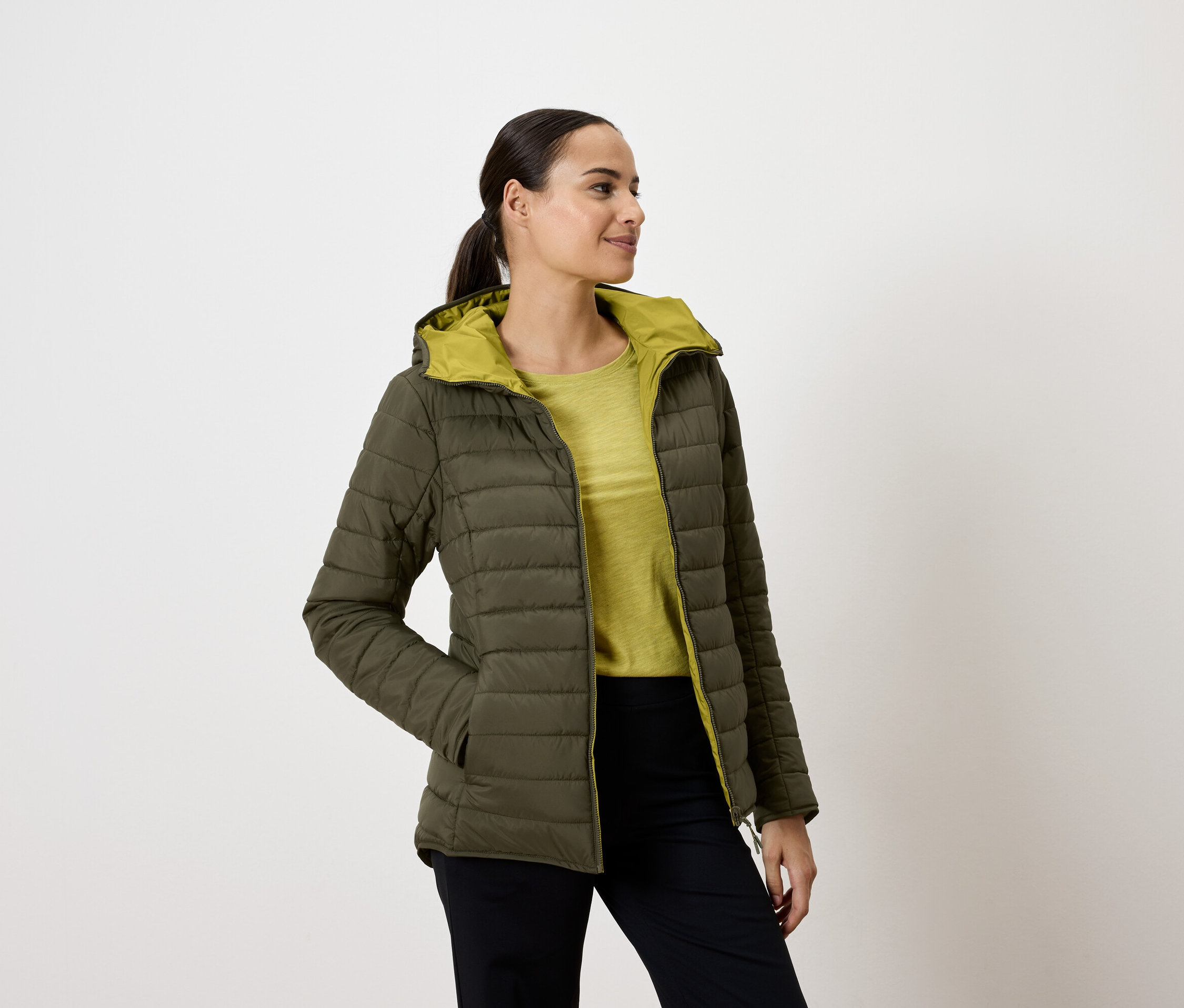 Nő olívazöld, steppelt kabátban, kapucnival, kezét a zsebében tartva, jobbra néz.