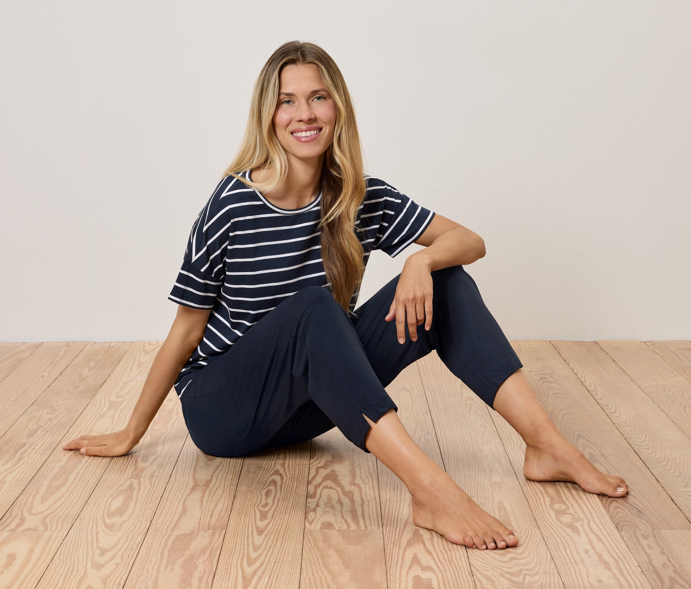 Egy szőke nő ül egy fapadlón, tengerészkék csíkos pólót és sötétkék nadrágot visel.