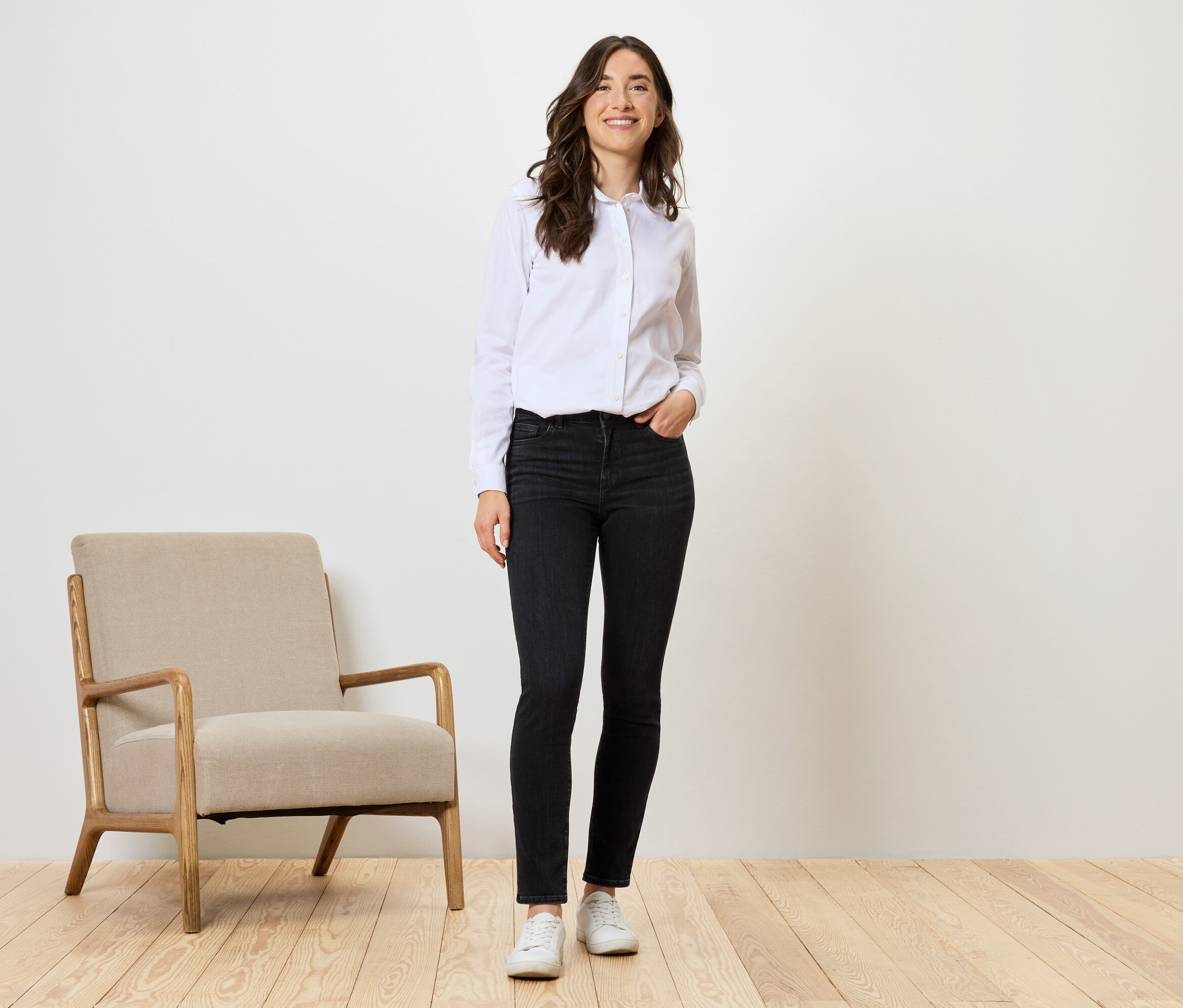 Egy nő áll fekete farmerben, fehér ingben és fehér cipőben egy bézs szék mellett.