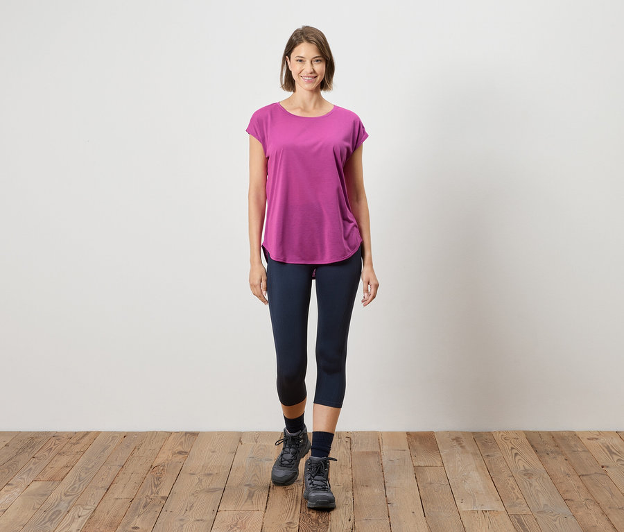 Egy nő áll egy fapadlón egy fehér fal előtt. Rózsaszín pólót, kék leggingset és szürke csizmát visel.