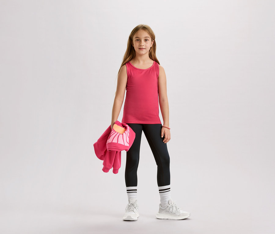 Lány rózsaszín trikóban, fekete leggingsben, fehér zokniban és tornacipőben, rózsaszín kabátot és táskát tart.