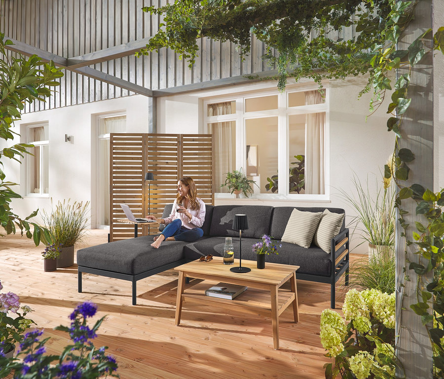 Egy nő ül a Prémium kerti sarokülőgarnitúrán, a háttérben egy rugalmas paraván látható. Látható még egy fekete LED akkumulátoros kültéri állólámpa és asztali lámpa, valamint egy kerti dohányzóasztal.