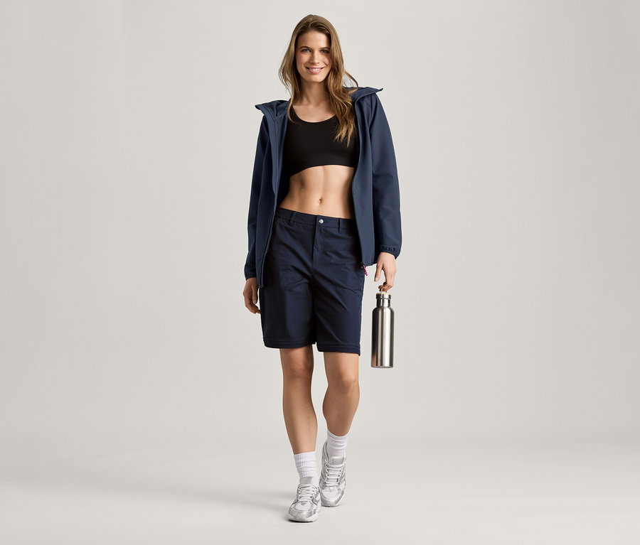 Egy nő sötétkék softshell dzsekiben, rövidnadrágban és sportmelltartóban ezüst színű kulacsot tart.