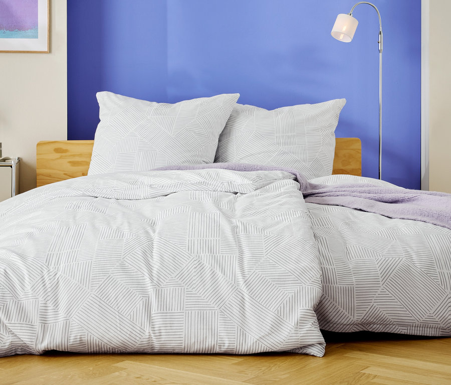 Egy ágy fehér ágyneművel, geometrikus mintával és két párnával, egy kék fal előtt.