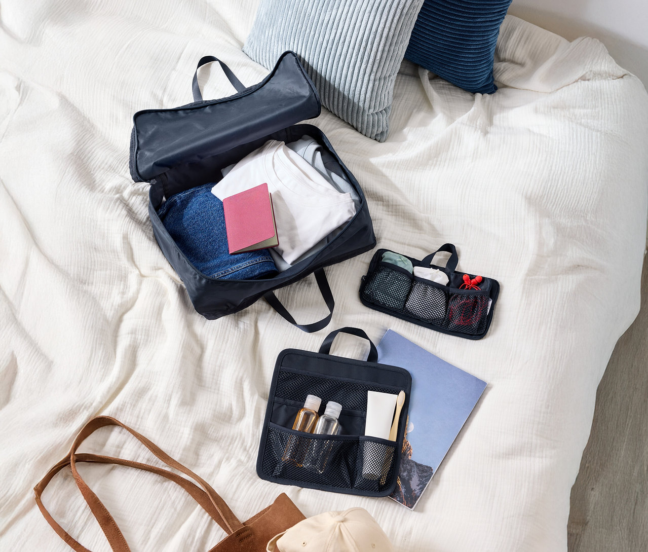 Felülnézet egy ruhákkal teli bőröndre, két párnára, egy kozmetikai táskára, egy tote táskára és egy sapkára egy ágyon.