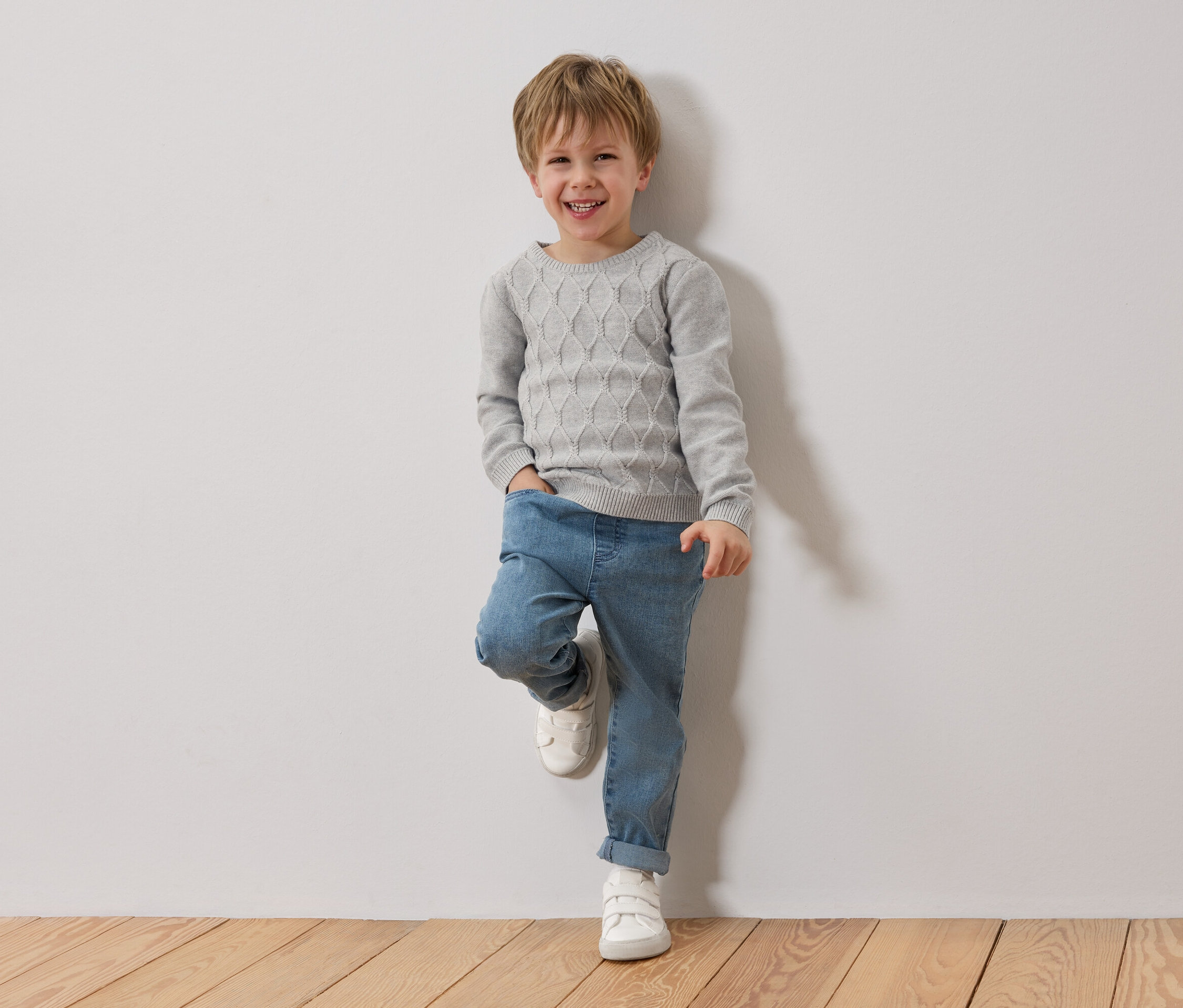 Egy mosolygós szőke fiú egy fehér falnak támaszkodik. Szürke pulóvert, kék farmert és fehér cipőt visel.