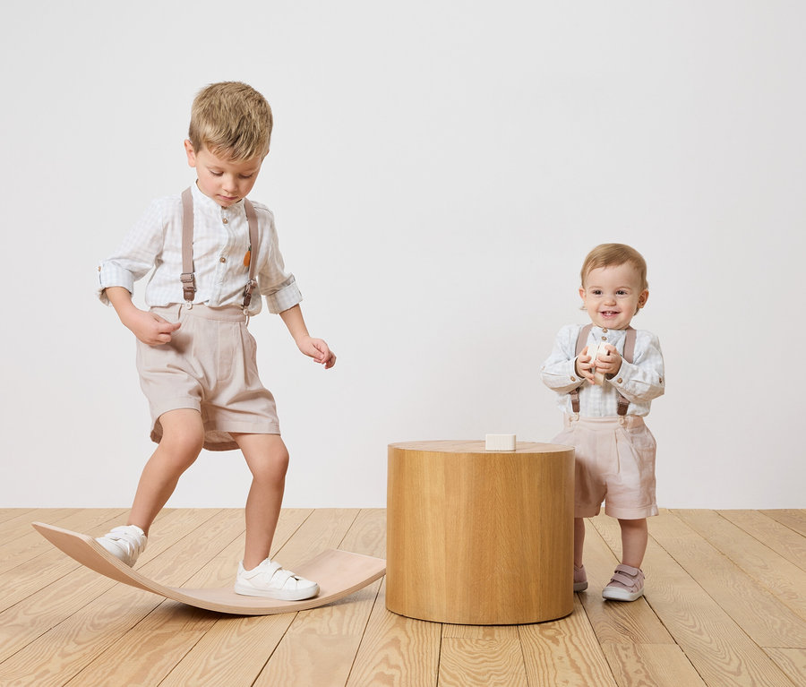 Két gyerek, egy fiú egyensúlyoz egy fa deszkán, egy csecsemő egy faasztal mellett áll és egy játékblokkot tart.