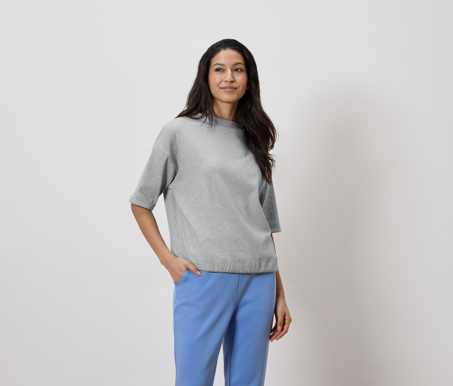 Sötét hajú nő szürke felsőt és kék nadrágot visel, keze a zsebében.